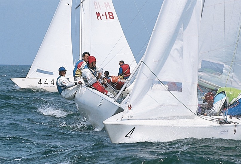 Das Boot im Vordergrund wendet gerade. Gegen den Wind laufend sind die Segel drucklos. Mit den Segeln wechselt auch die Crew die Seite.  Kurz zuvor lief das Boot auf Backbordbug (wie das Boot dahinter). Nach der Wende läuft es auf Steuerbordbug (wie das Boot 444 im Hintergrund) weiter.